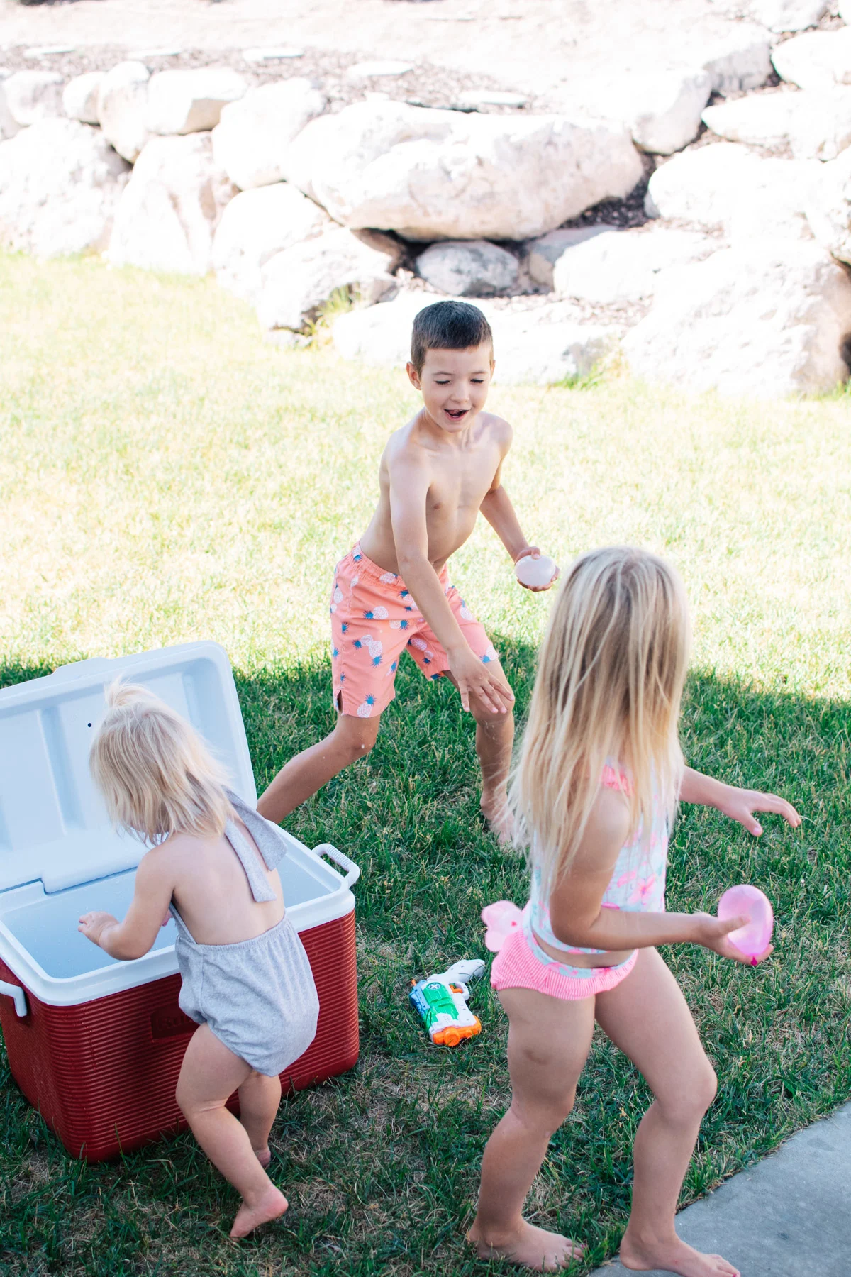Boy throws water balloon at young girl while baby gets a balloon out of red cooler.