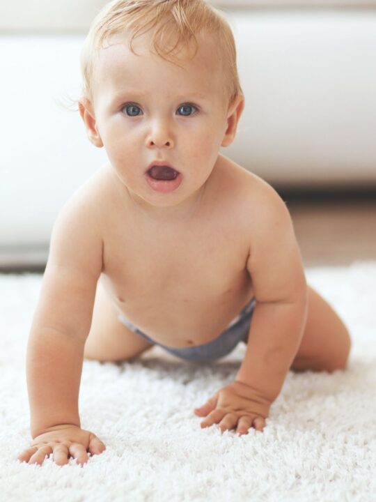 Baby boy with strawberry blonde hair and blue eyes crawls on white rug.