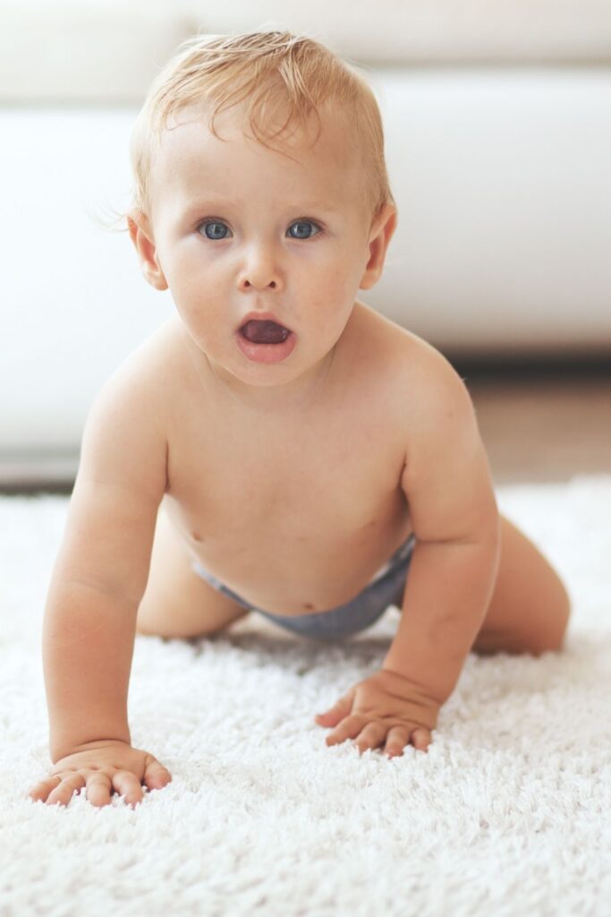 Baby boy with strawberry blonde hair and blue eyes crawls on white rug.