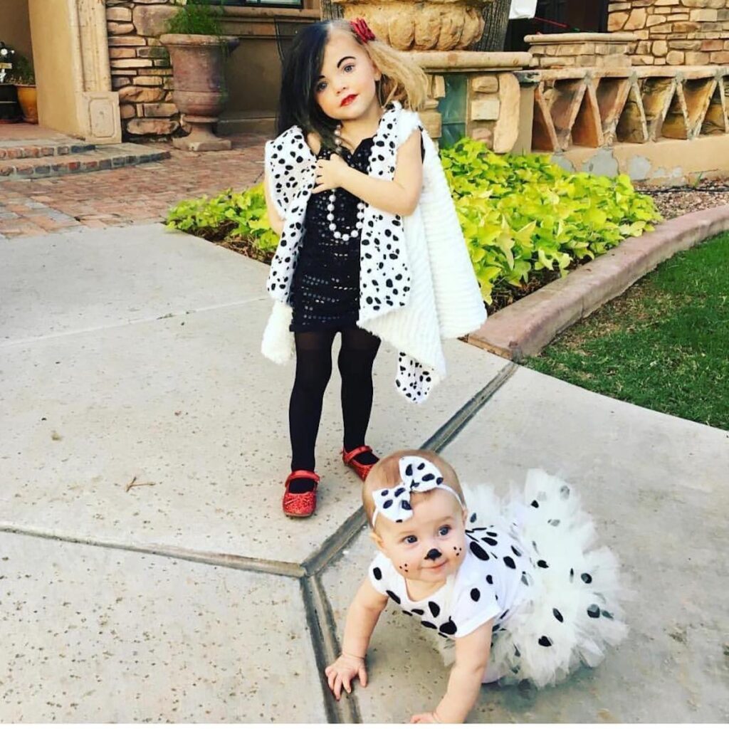 Toddler girl dressed as Cruella DeVil stands behind baby dresses as dalmatian.