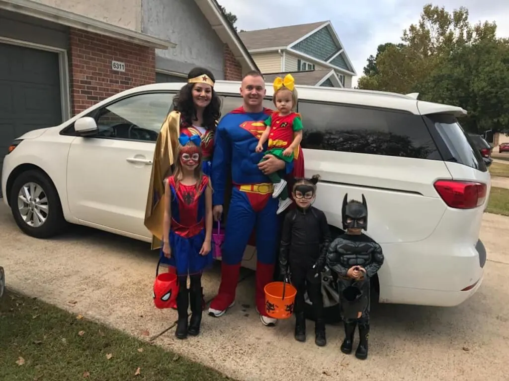 Family wearing superhero costumes smiles for picture in front of white van.