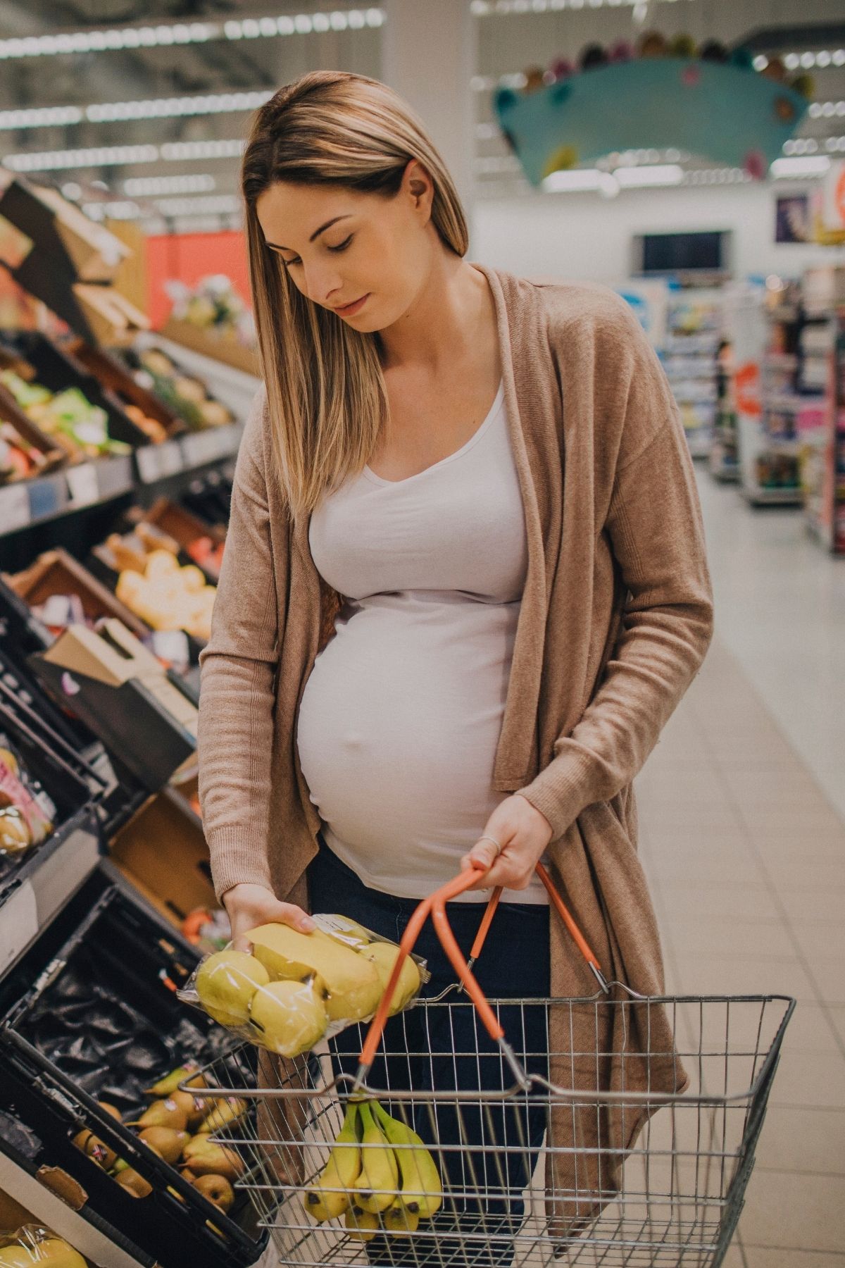 Pregnant woman in grocery store holds bag of apples over her shopping basket carrying bananas.