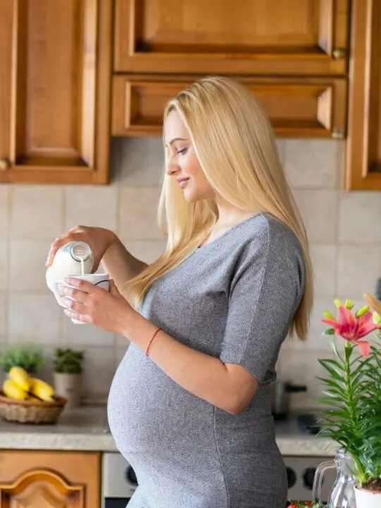 Pregnant woman wearing gray dress pours milk from a glass jar into a glass cup.