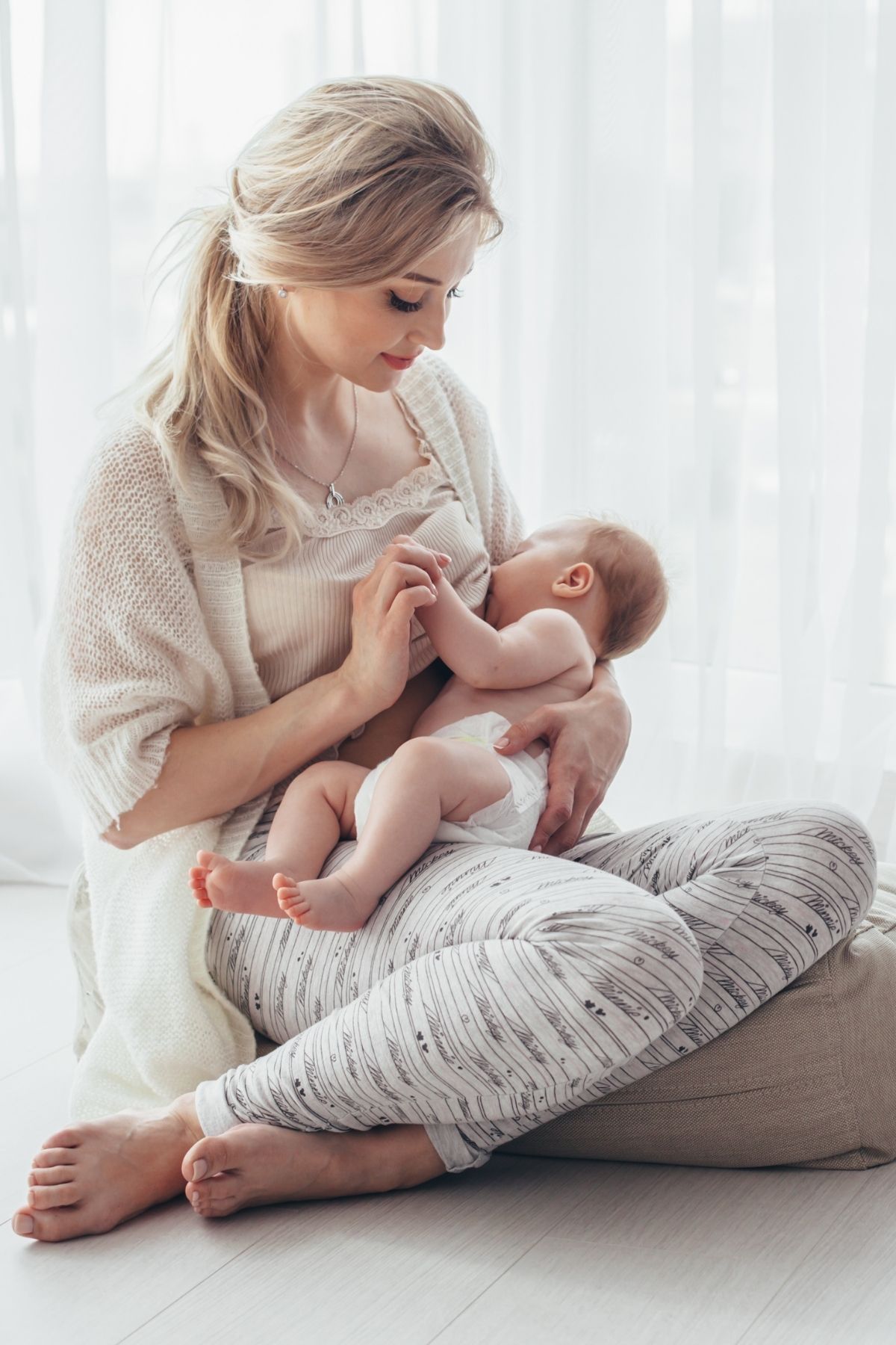 Blonde woman nurses her baby while sitting on a bean bag in front of window.