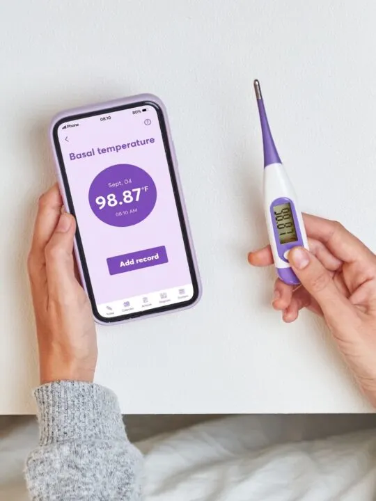 Woman holds basal thermometer and app to record a rise in BBT after ovulation.