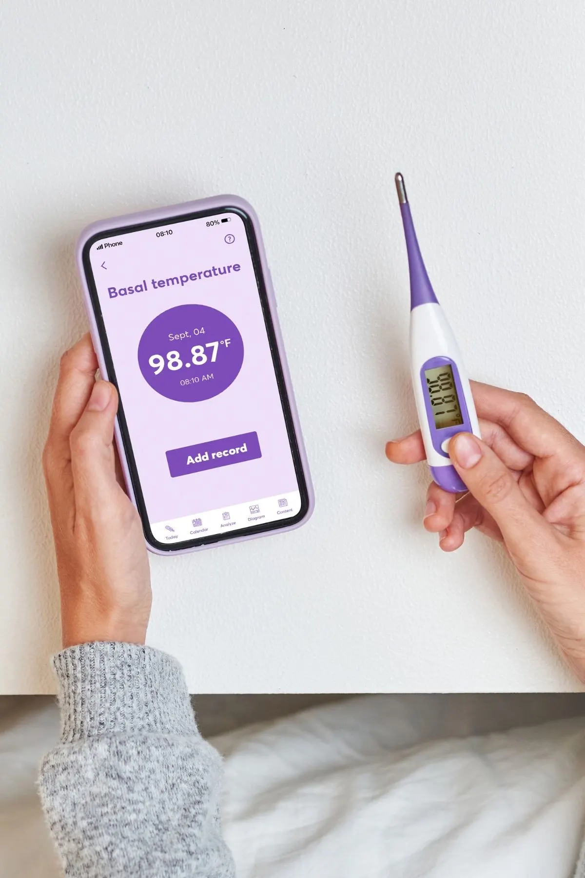 Woman holds purple basal thermometer and app to record a rise in BBT after ovulation.