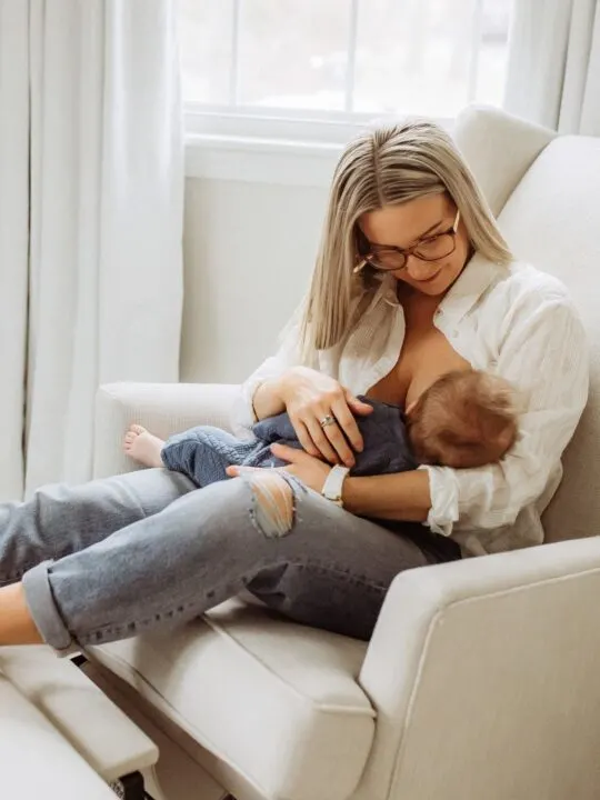 New mom breastfeeds baby boy while sitting on glider in nursery room.