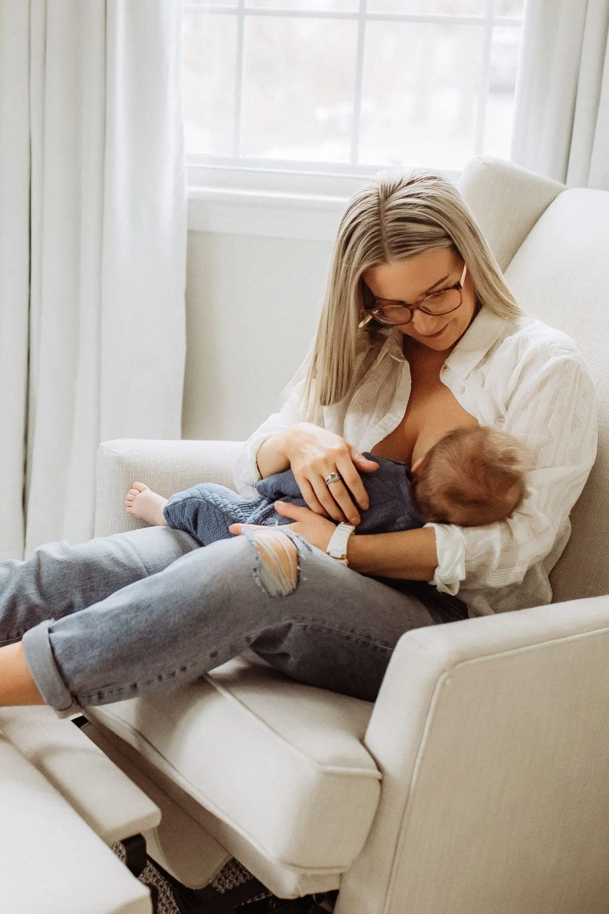 Woman with blonde hair breastfeeds baby boy while sitting on glider in nursery room.