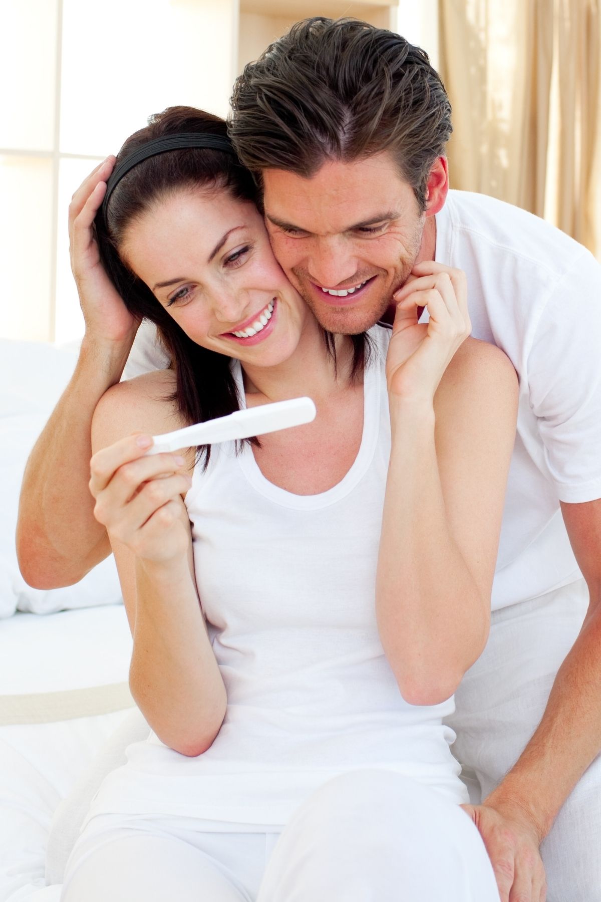 A couple wearing white shirts embraces as they look at the results of a pregnancy test.