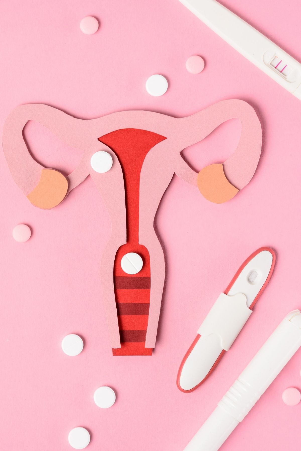 Paper diagram of uterus surrounded by white and pink pills and several pregnancy tests on pink background.