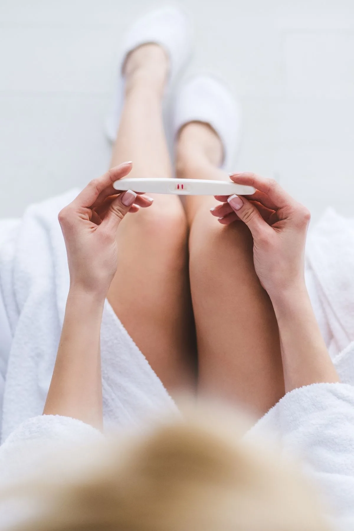 Woman wearing white robe and slippers holds positive pregnancy test above her knees.