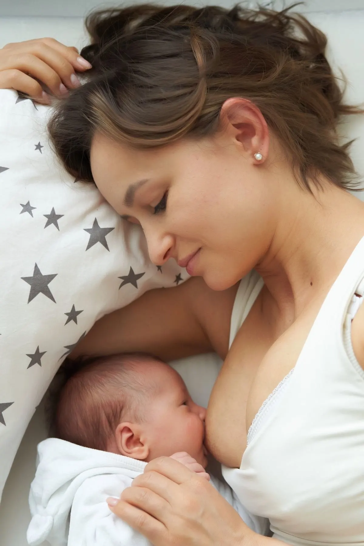 Brunette woman breastfeeds baby while laying down on a white nursing pillow with gray stars.
