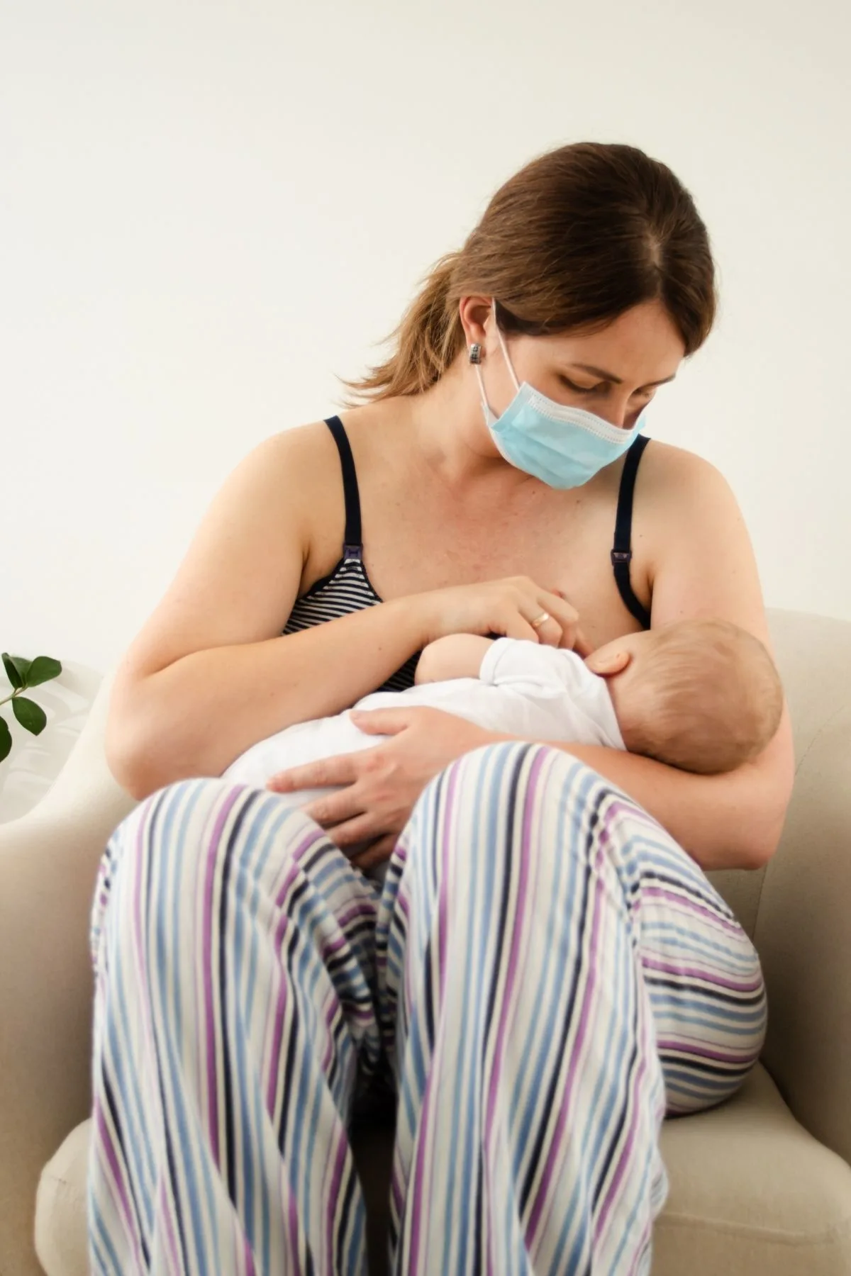 Brunette woman wearing face mask and pajamas sits in beige chair and nurses baby.