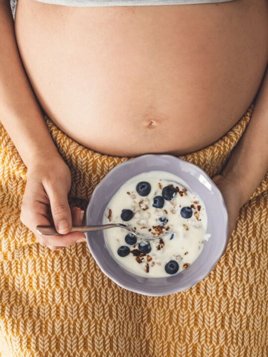 Pregnant woman with belly exposed eats a bowl of cereal with blueberries.
