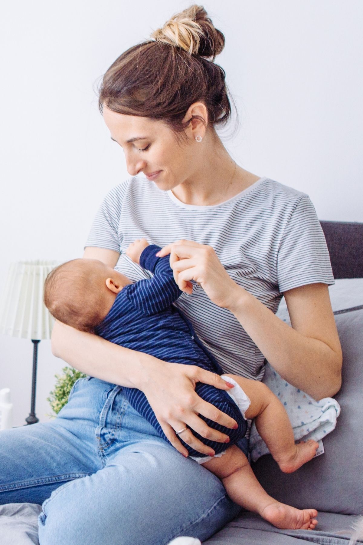 Woman in gray shirt sits on bed to breastfeed her son in blue onesie.