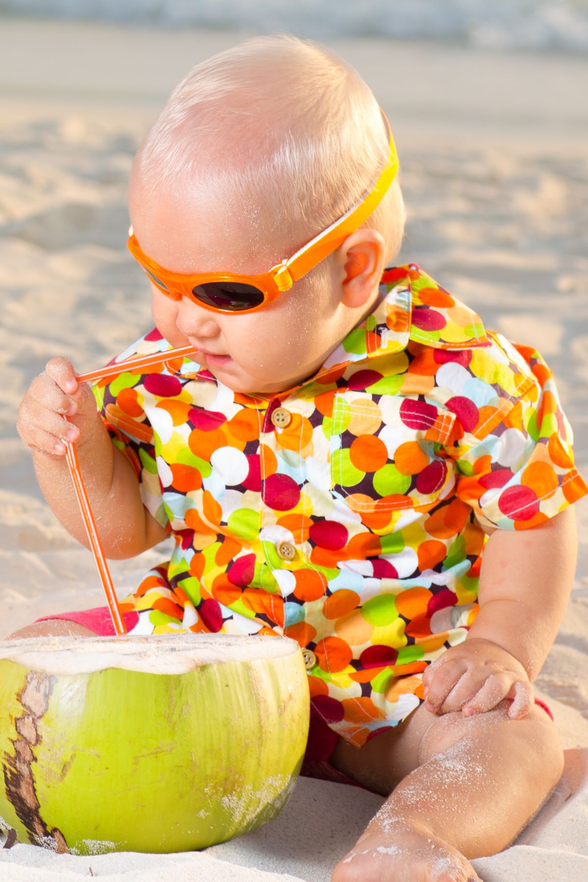 Baby boy in sunglasses and colorful clothes drinks from a green coconut on a beach.