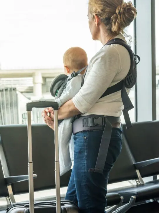 Mom holds her baby in a carrier with her luggage in front of airport window.
