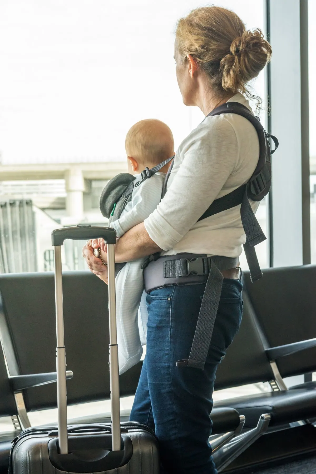 Mom holds her baby in a carrier with her luggage in front of airport window.