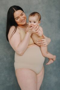New mom in nude undergarments holds her baby while standing in front of gray background.