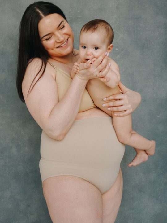 New mom in nude undergarments holds her baby while standing in front of gray background.