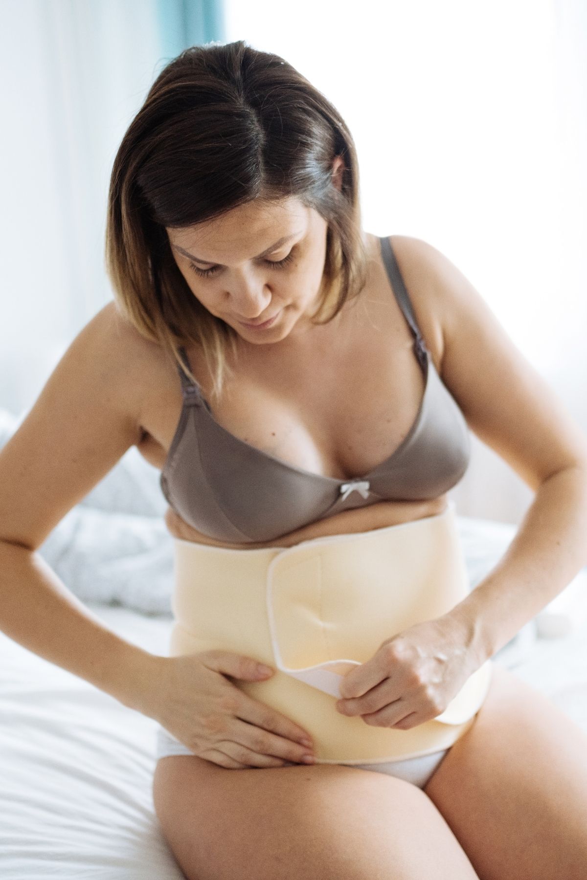 New mom sits on her bed while putting on a postpartum girdle.