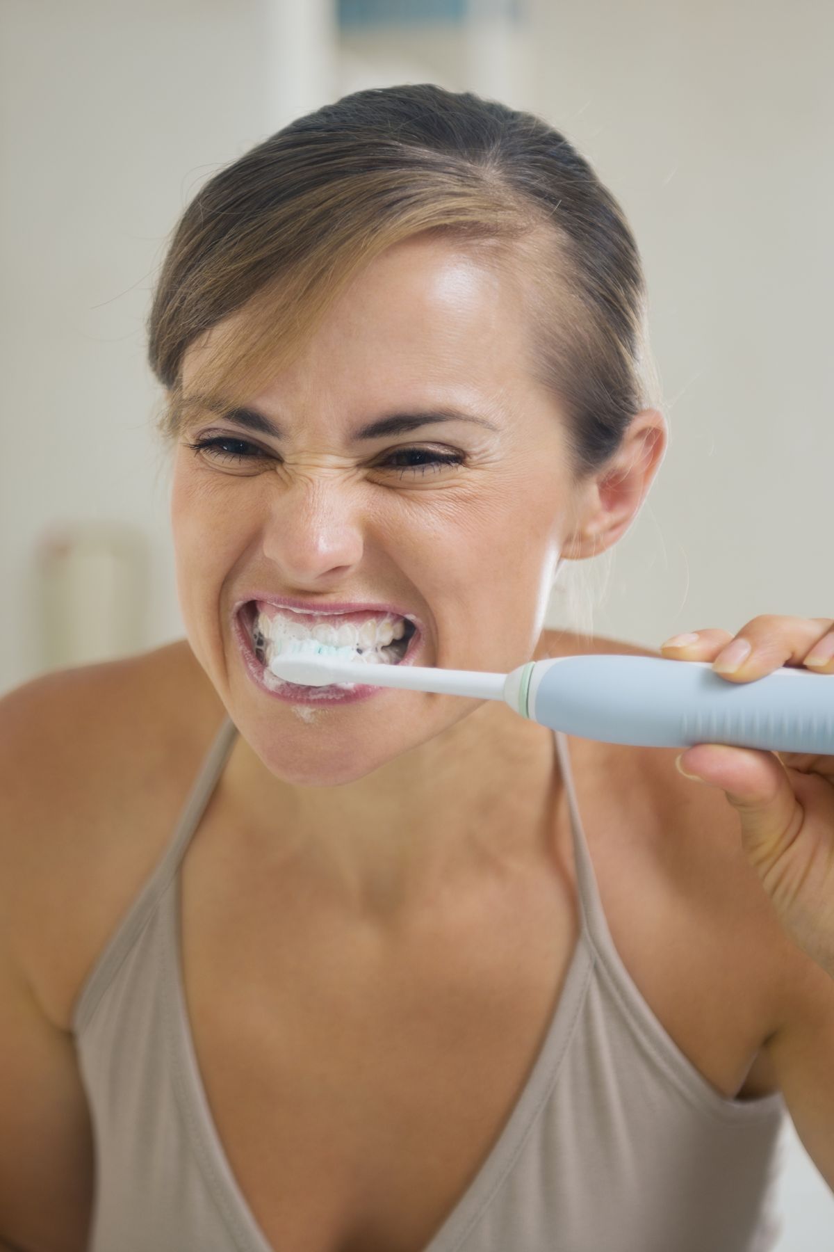 Woman in gray tank top brushes her teeth with an electric toothbrush.
