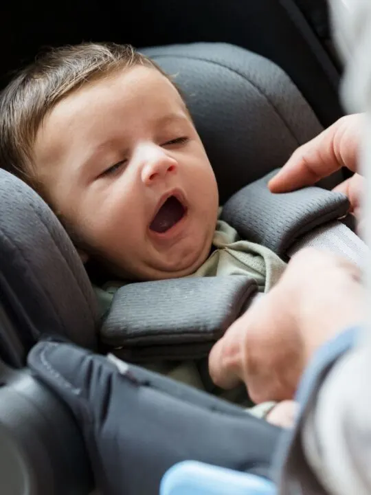 A parent straps a yawning baby into a gray infant car seat.