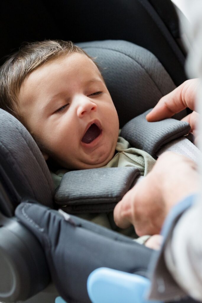 A parent straps a yawning baby into a gray infant car seat.
