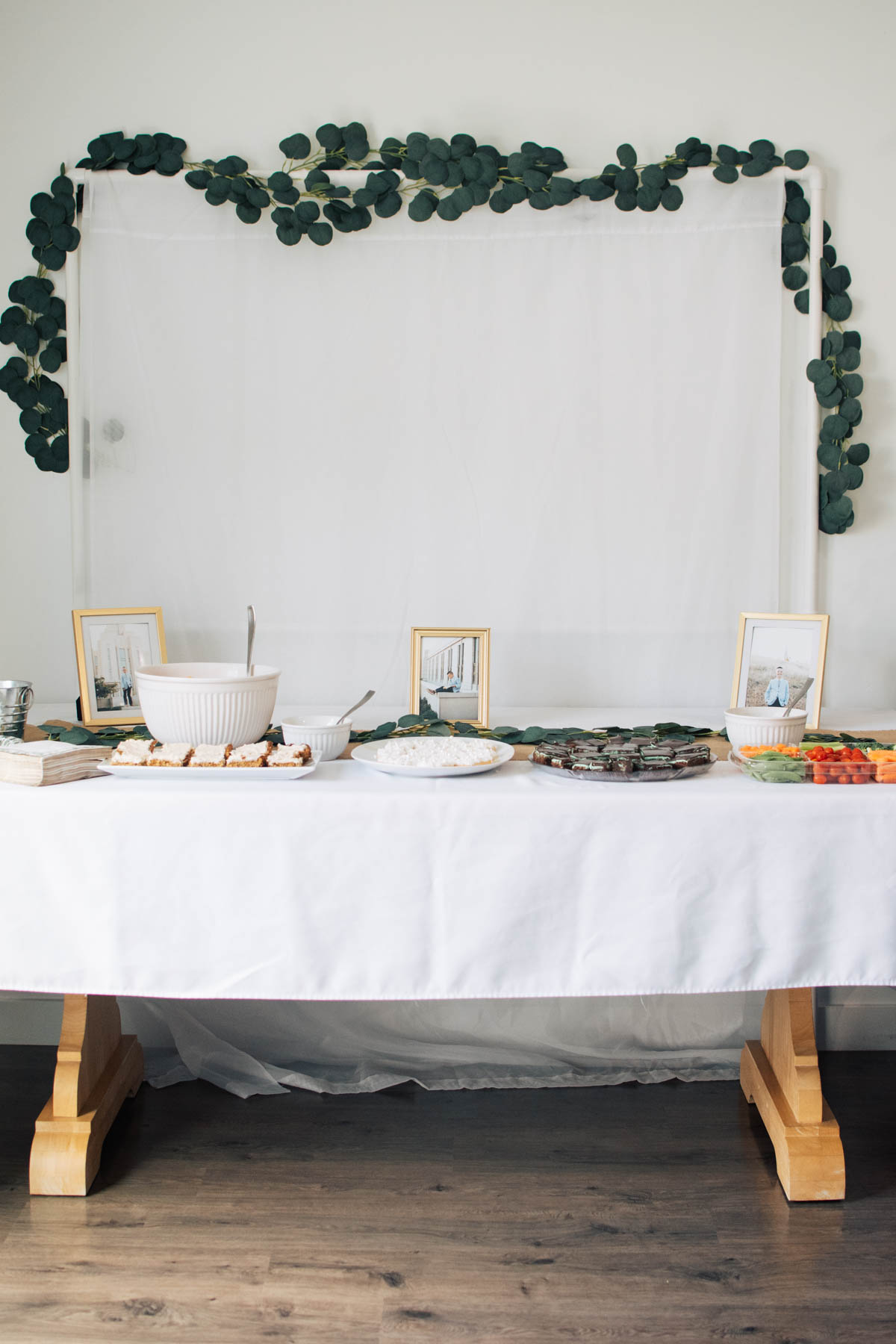 LDS baptism party backdrop with eucalyptus garland and food table with white table cloth.