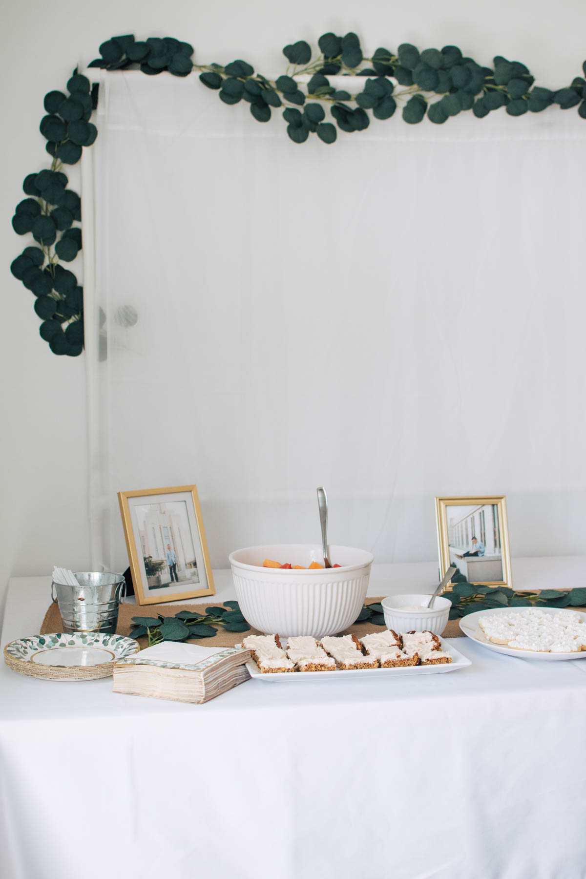 LDS baptism party backdrop and food table with eucalyptus garland and white table cloth.