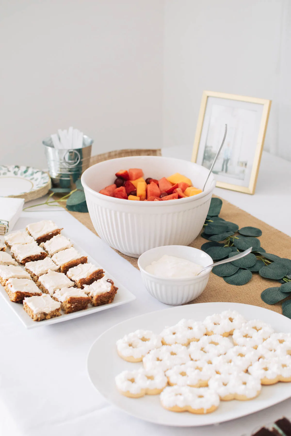 Plates of banana bars, cookies, and bowl of fruit on party food table.