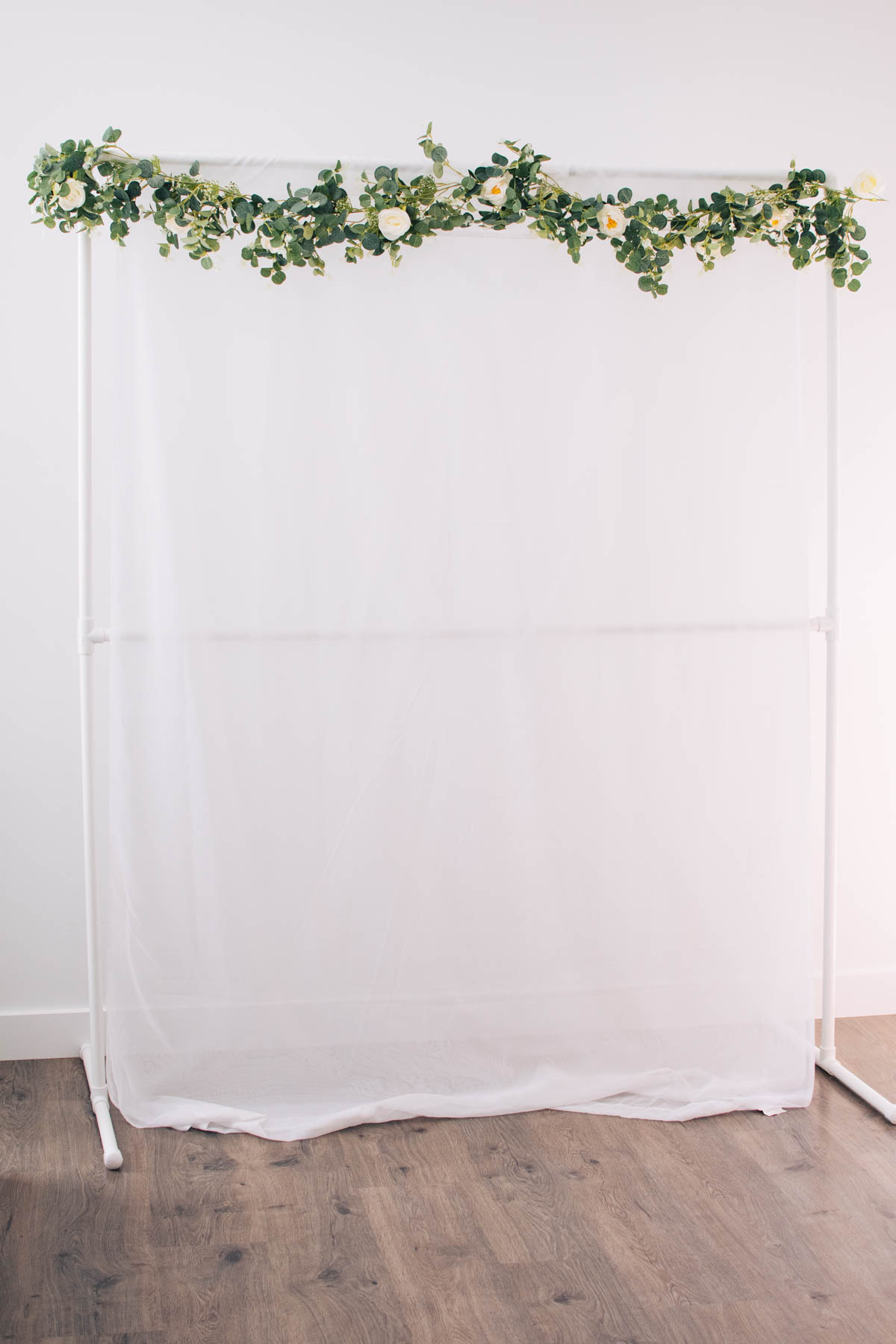 PVC backdrop stand with sheer curtain and floral garland draped along the top in empty room.