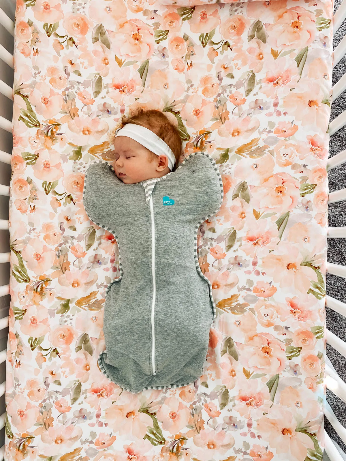 Baby wearing a zipper swaddle sleeps in white crib with peach floral sheet.