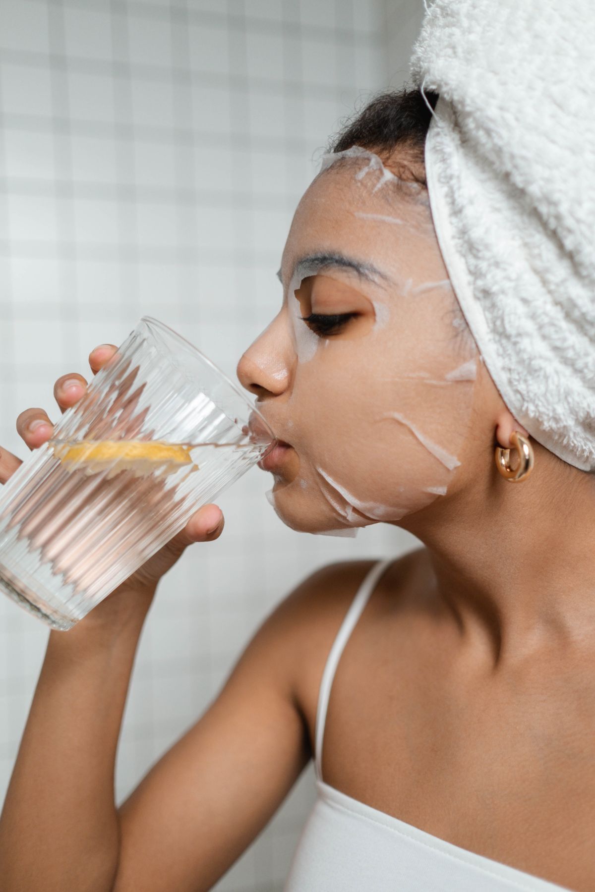 Woman drinking a glass of water with a skin care face mask and hair wrapped in a towel.