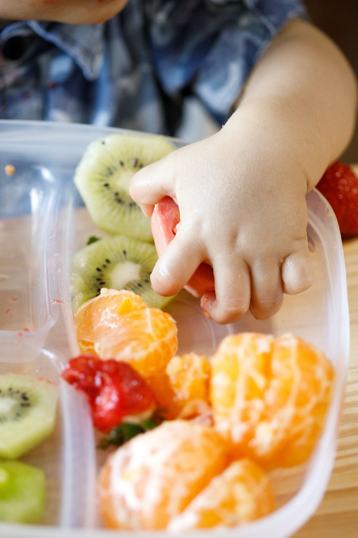 A toddler's hand grabs a piece of fruit from a plastic tray of assorted fruits.