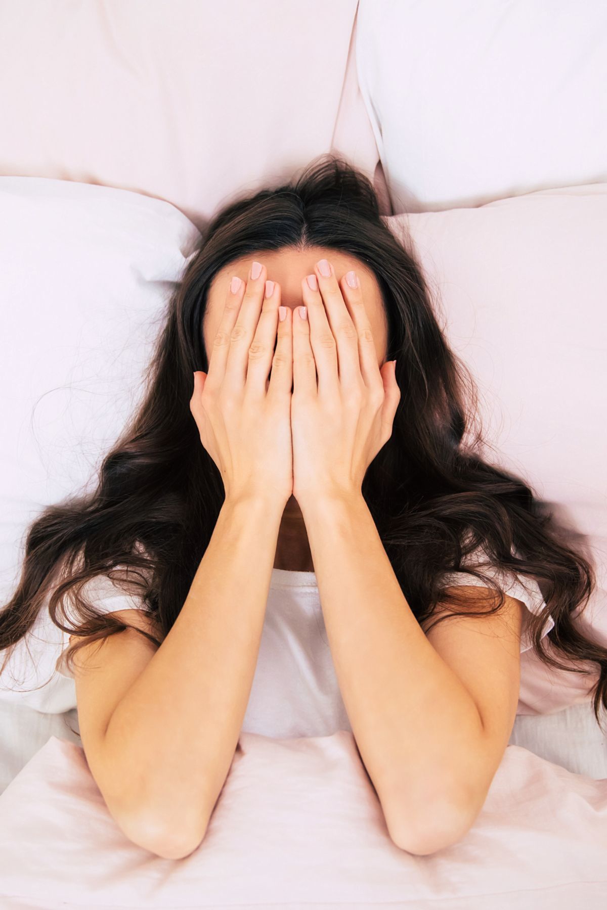 A woman with long dark hair lays in bed with her hands over her face.