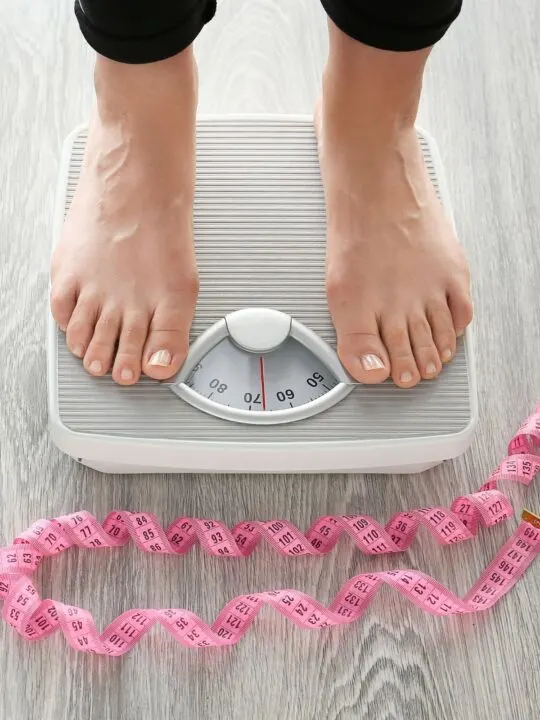 A woman stands on a gray and white bathroom scale next to pink measuring tape.