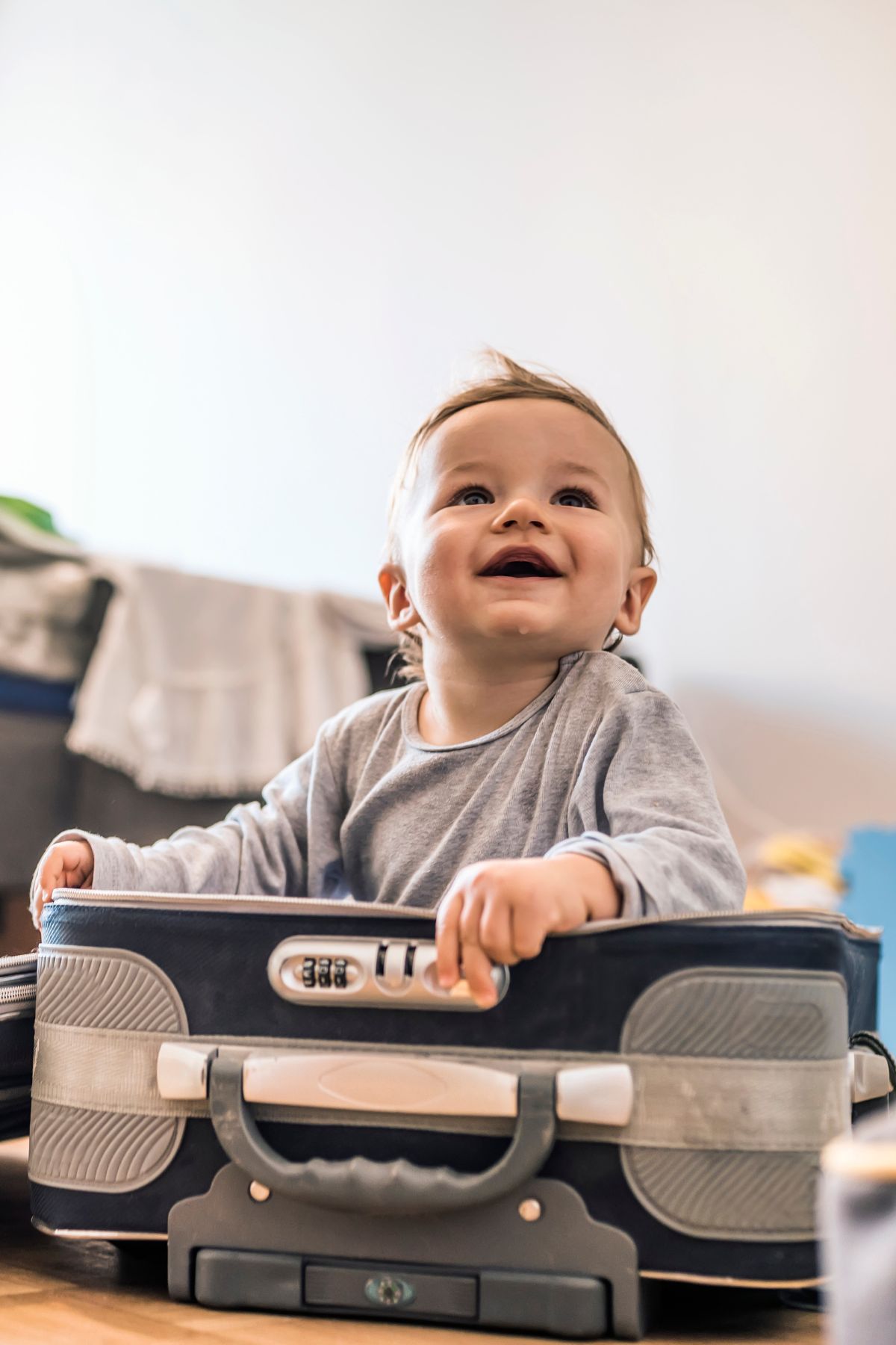 A smiling baby boy sits in an unpacked suitcase in a living room.