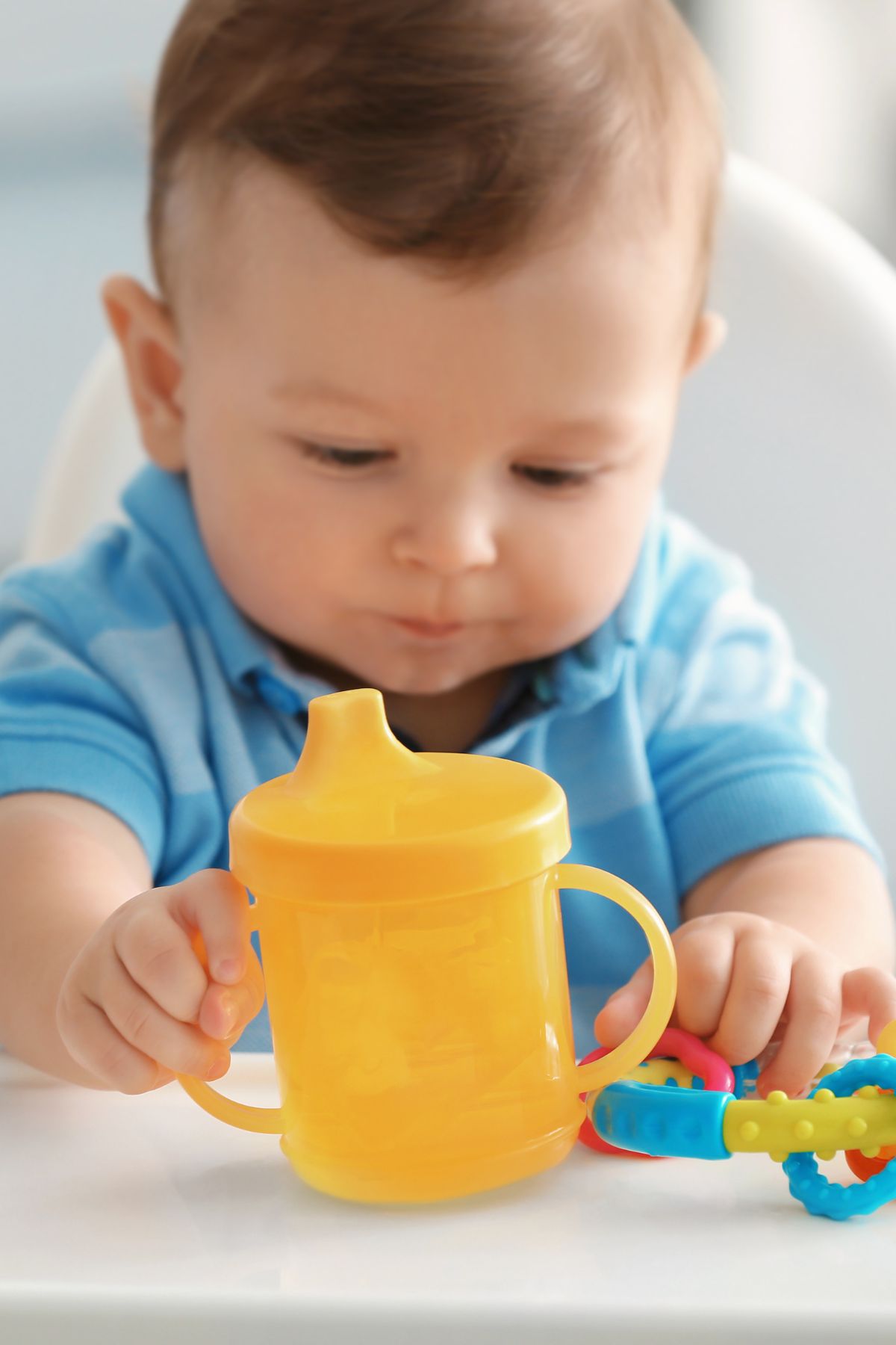 A toddler boy holds a yellow sippy cup while sitting in a high chair.