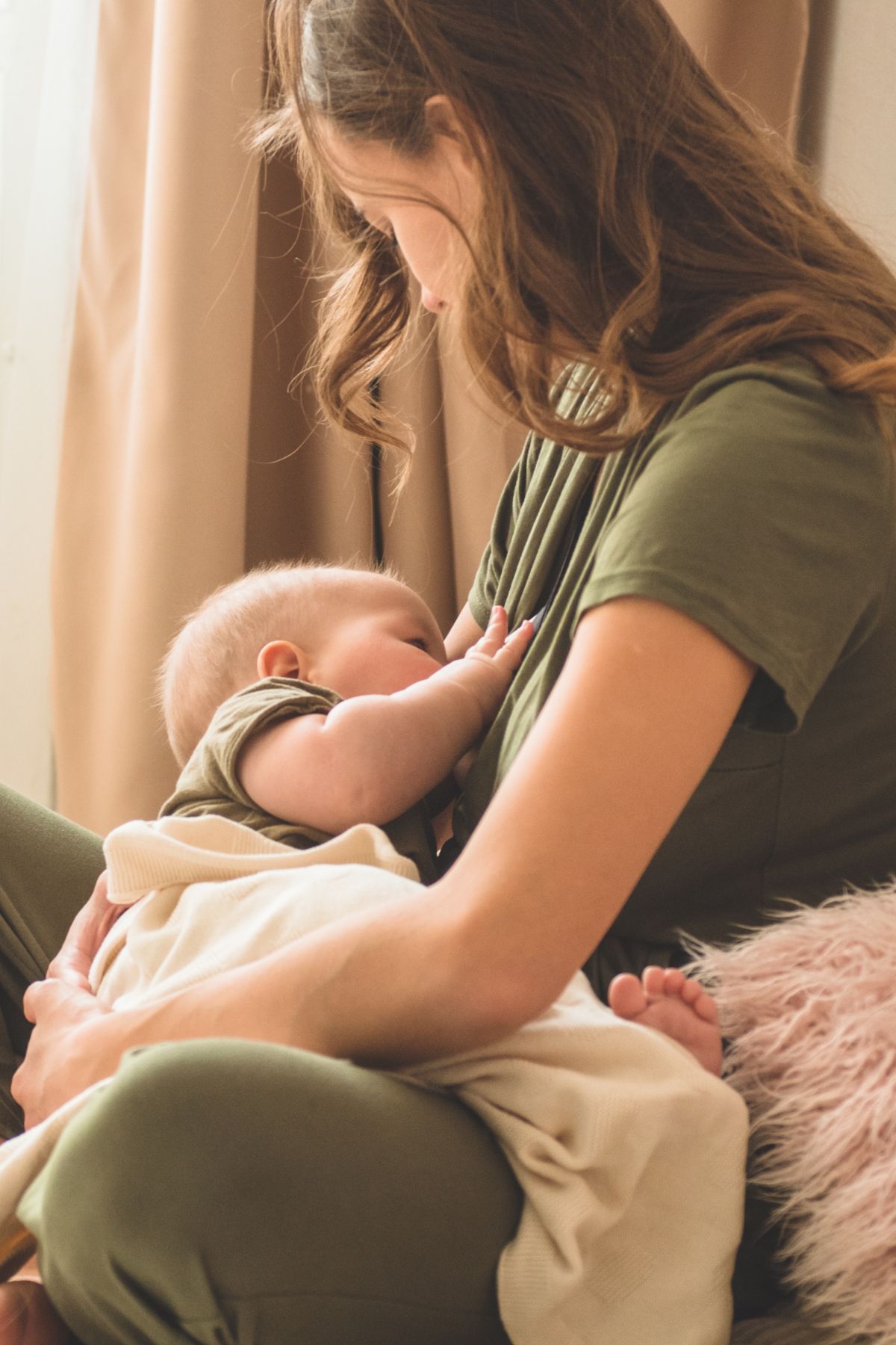 A woman in a green shirt and pants breastfeeding her baby on a bed.