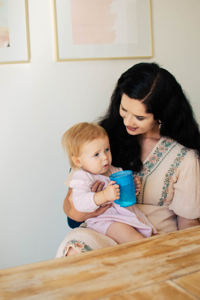 Brunette woman with baby girl in pink dress on her lap; the baby is holding a blue sippy cup with milk.