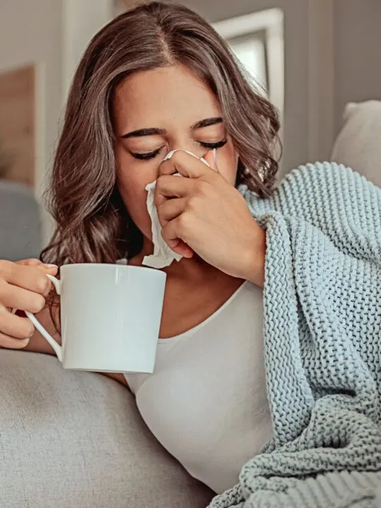 A sick woman blows her nose while holding a white mug.