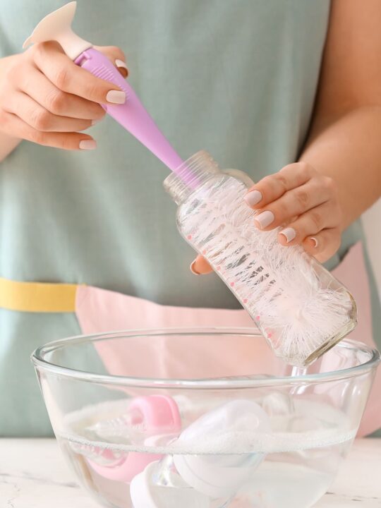 A woman sterilizes baby gear in a clear glass bowl using a bottle brush.