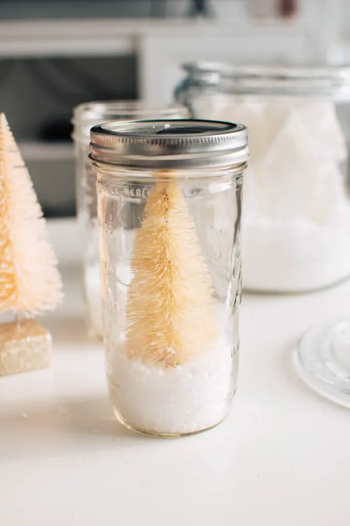 Cream bottle brush tree and fake snow in Mason jar on white table.