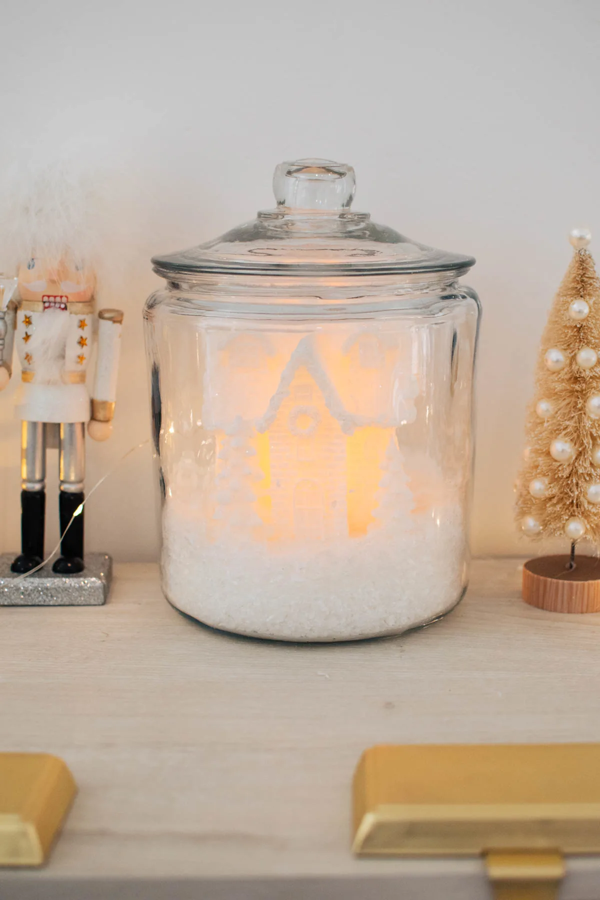 White Christmas house with light and fake snow in glass cookie jar on cream cabinet.