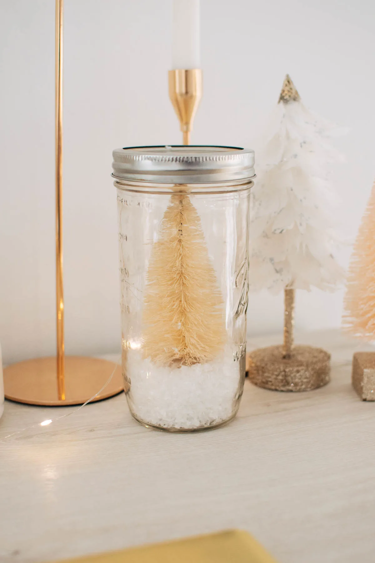 Cream bottle brush tree and fake snow in Mason jar next to gold candlesticks on cream cabinet.