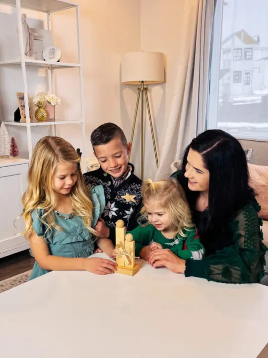 Mom with kids sits around a white table with small wood nativity scene on it.