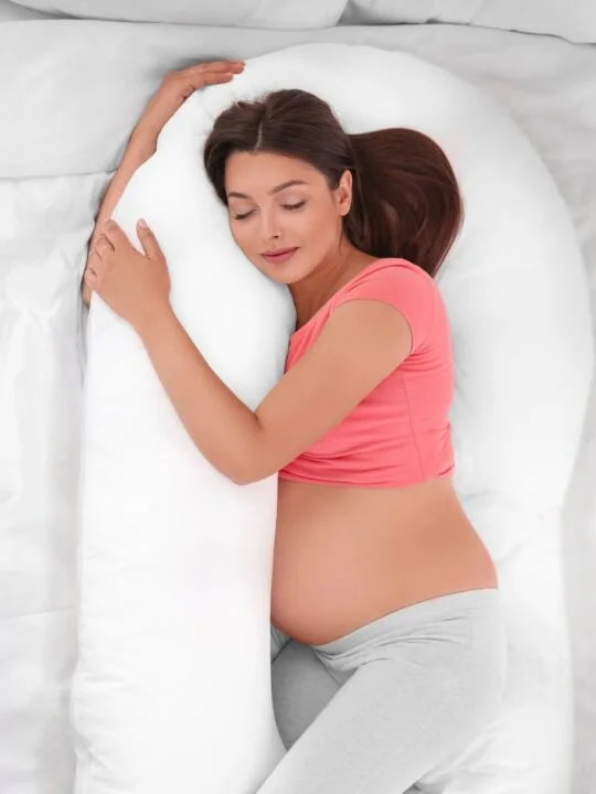A pregnant woman sleeps with a large white body pillow on a bed.