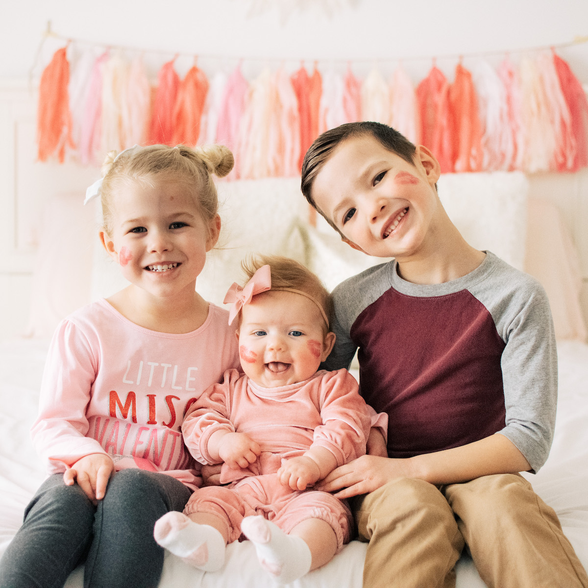 3 Children pose with lipstick kisses on their faces in front of a pink garland.