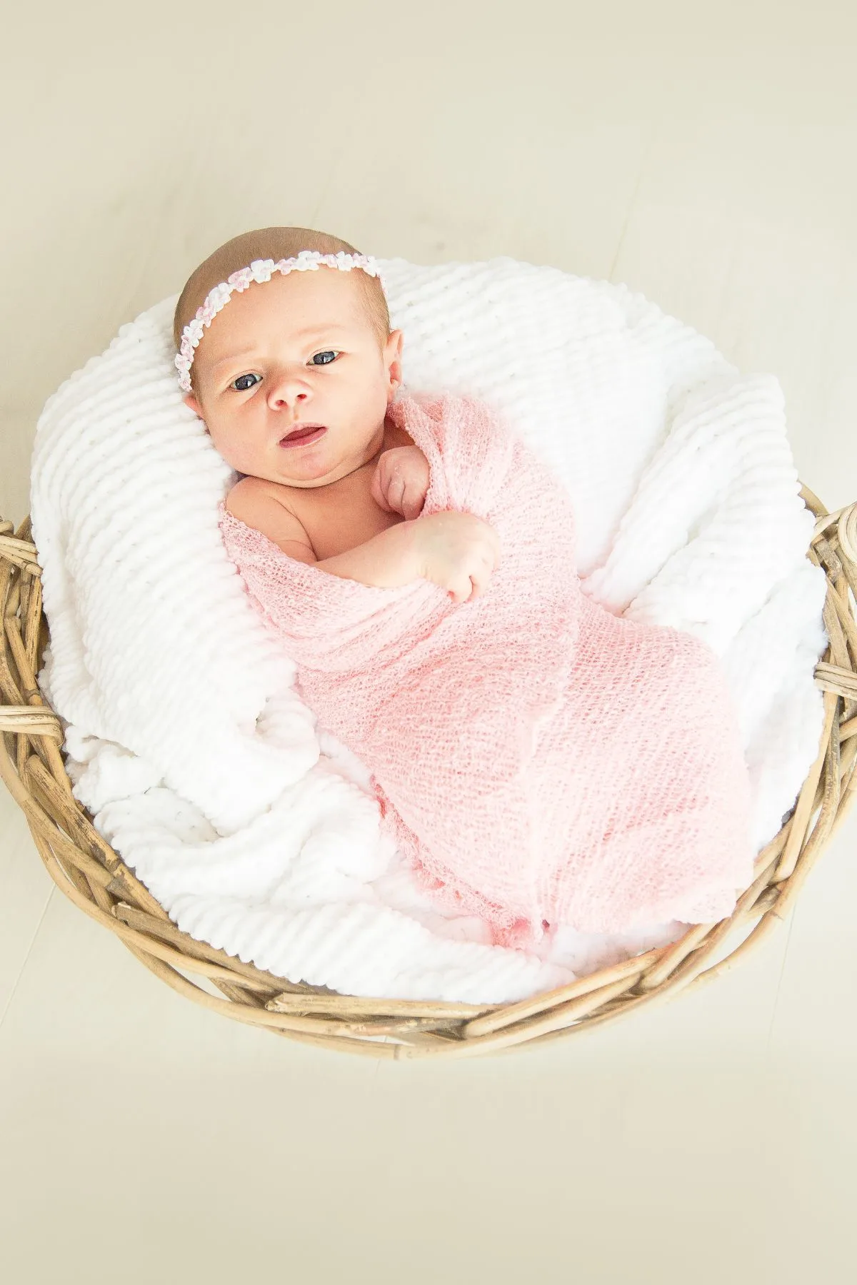 Baby girl in pink swaddle and headband resting on white blankets in wicker basket.